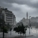 Πλατεία στο κέντρο της Αθήνας με ορατό τον Πύργο του Αγίου Κωνσταντίνου στο βάθος, βυθισμένη σε σφοδρή νεροποντή. Οι δρόμοι έχουν μετατραπεί σε λίμνες, τα δέντρα λυγίζουν από τον αέρα, τα φανάρια και τα αυτοκίνητα αντανακλούν στα νερά και ο ουρανός είναι σκοτεινός και μολυβένιος. Σκηνή ακραίων καιρικών φαινομένων στην Αθήνα.