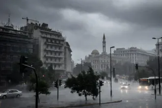 Πλατεία στο κέντρο της Αθήνας με ορατό τον Πύργο του Αγίου Κωνσταντίνου στο βάθος, βυθισμένη σε σφοδρή νεροποντή. Οι δρόμοι έχουν μετατραπεί σε λίμνες, τα δέντρα λυγίζουν από τον αέρα, τα φανάρια και τα αυτοκίνητα αντανακλούν στα νερά και ο ουρανός είναι σκοτεινός και μολυβένιος. Σκηνή ακραίων καιρικών φαινομένων στην Αθήνα.