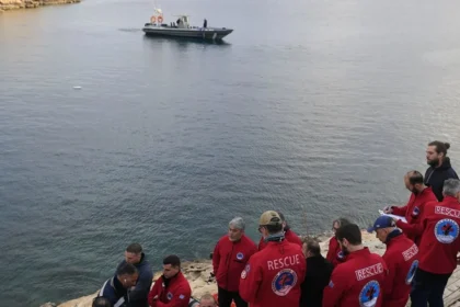 Άνδρες των σωστικών συνεργείων επιχειρούν σε βραχώδη ακτή με φόντο τη θάλασσα, γύρω από σωστικό εξοπλισμό.