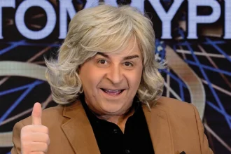 O Μάρκος Σεφερλής με θεατρικό κοστούμι σε σκηνή με έντονο φωτισμό εκτελεί αστείες κινήσεις.