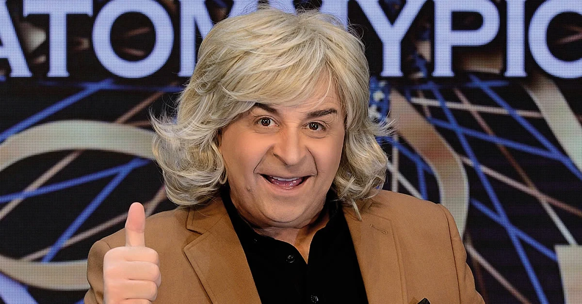 O Μάρκος Σεφερλής με θεατρικό κοστούμι σε σκηνή με έντονο φωτισμό εκτελεί αστείες κινήσεις.