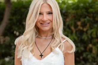 Η Φαίη Σκορδά ποζάρει λαμπερή, φορώντας ένα κομψό λευκό μίνι φόρεμα με λεπτομέρειες δαντέλας και εντυπωσιακά χρυσά ψηλοτάκουνα πέδιλα.