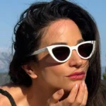 Η Κατερίνα Γερονικολού ποζάρει δίπλα στην πισίνα φοράει μαύρο μπικίνι, λευκά γυαλιά cat-eye και visor καπέλο.