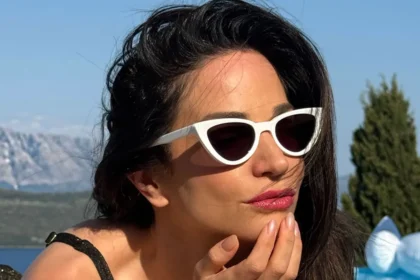 Η Κατερίνα Γερονικολού ποζάρει δίπλα στην πισίνα φοράει μαύρο μπικίνι, λευκά γυαλιά cat-eye και visor καπέλο.