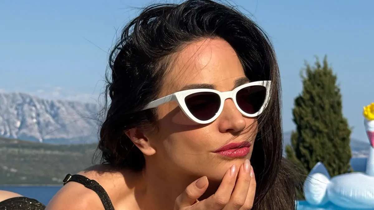 Η Κατερίνα Γερονικολού ποζάρει δίπλα στην πισίνα φοράει μαύρο μπικίνι, λευκά γυαλιά cat-eye και visor καπέλο.